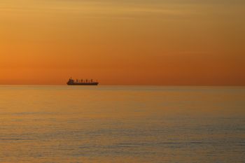 A summer sunrise over a calm Baltic Sea with the silhouette of a ship on the horizon colours the water and the sky deeply red.